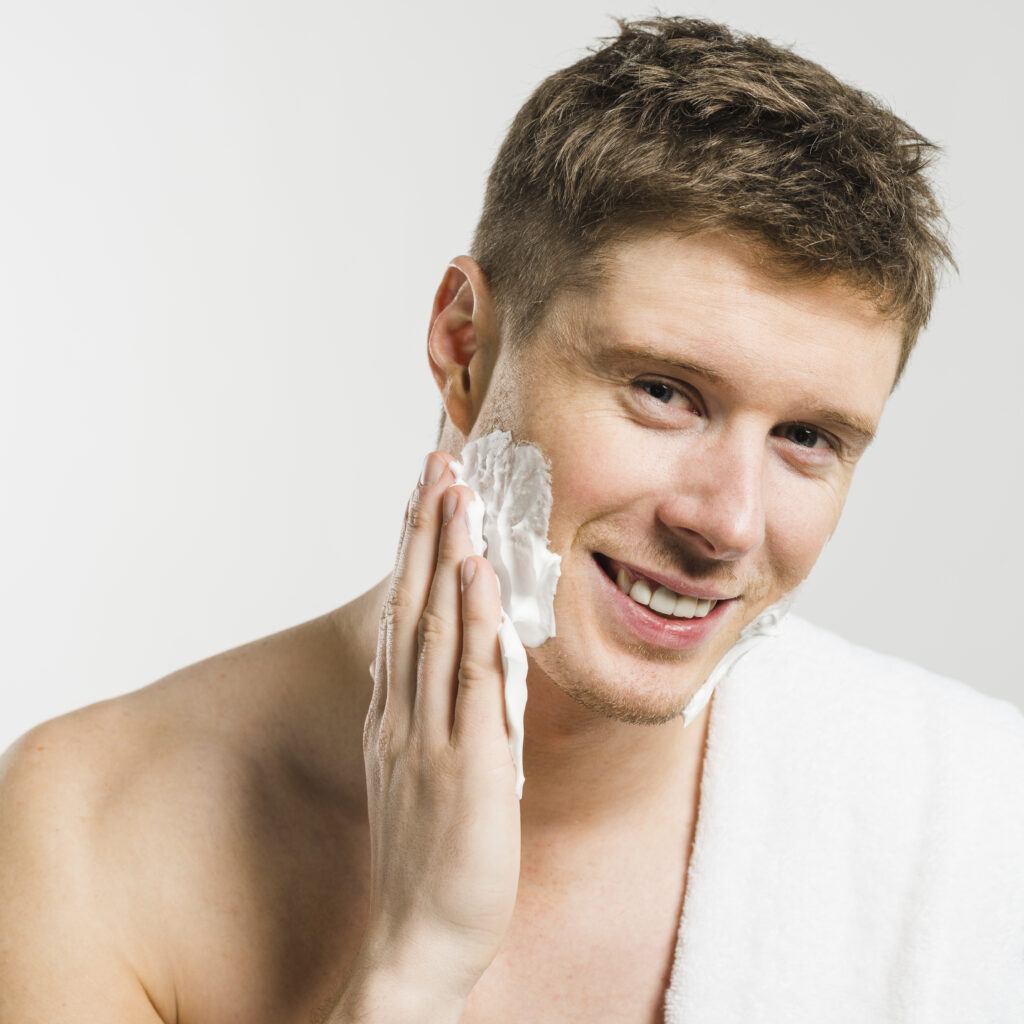 portrait smiling man applying shaving foam his face with hand against white backdrop
